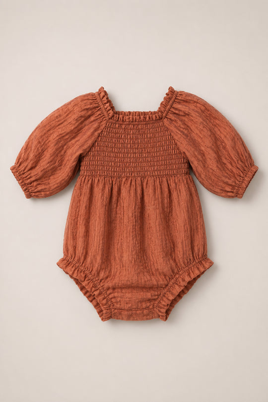 Ivy City Co’s Baby Madeline Romper in Apple Cider features a rust-colored long-sleeve design with a smocked bodice, subtle puff sleeves, elastic cuffs, and gathered leg openings, displayed on a neutral background.