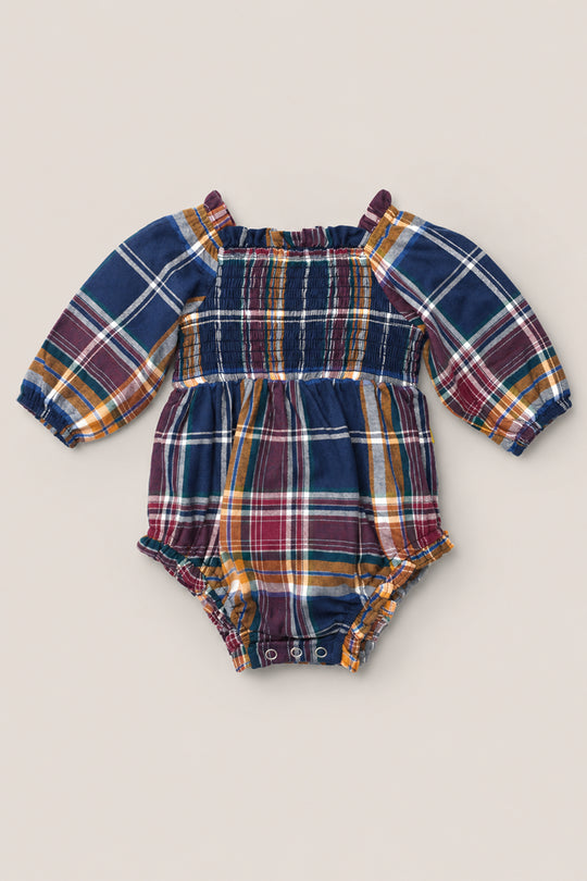 The Ivy City Co Baby Madeline Romper in Midnight Plaid features long sleeves, a vibrant blue, red, yellow, and white plaid pattern, a smocked bodice for extra charm, and gathered elastic at the chest and leg openings.