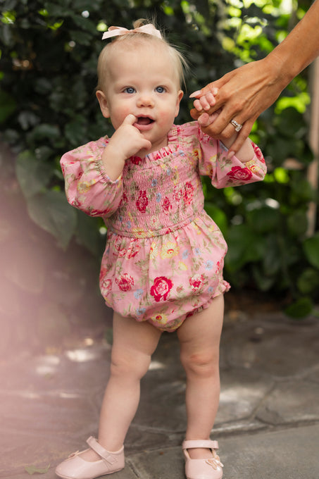 A baby in the Ivy City Co Baby Madeline Romper in Pink Vintage Poppies stands on a stone path, holding an adult's hand for support, surrounded by lush greenery.