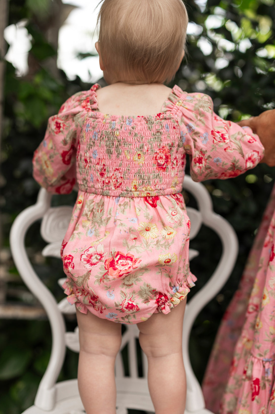 A light-haired baby stands on a white chair, wearing the Ivy City Co Baby Madeline Romper in Pink Vintage Poppies with puffed sleeves, ruffles, and a smocked bodice. Greenery is visible in the background.