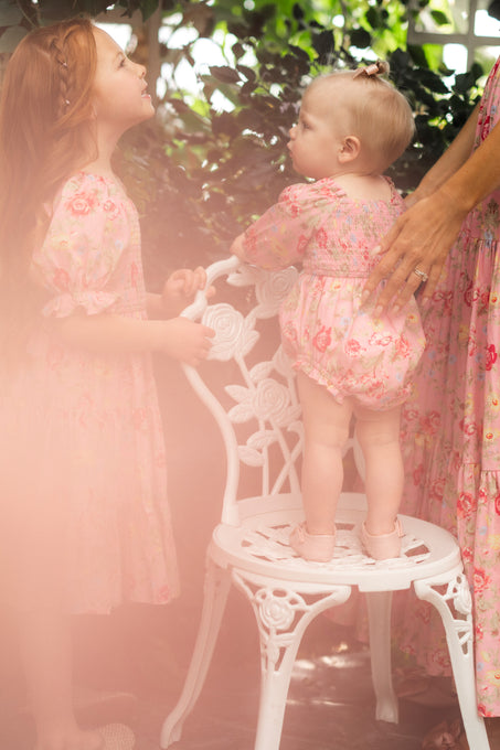 Two young children wear Ivy City Co’s Baby Madeline Romper in Pink Vintage Poppies; the older girl faces the baby, who stands on a white decorative chair with gentle adult support and dreamy soft pink lighting.