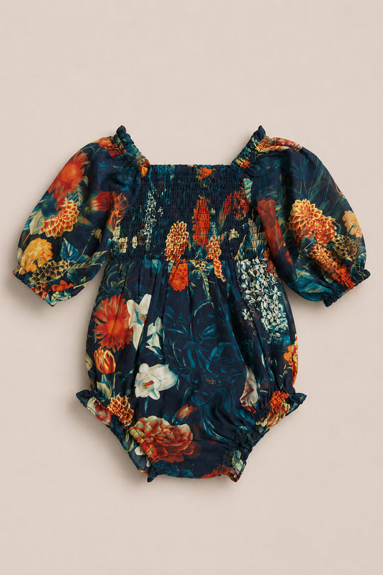 The Baby Madeline Romper in Imogen Moody Floral by Ivy City Co features a smocked bodice with charming puff sleeves, set against a dark blue background adorned with a colorful floral pattern of orange, red, and white flowers. This adorable chiffon romper also includes elastic leg openings for comfort and is beautifully displayed on a plain beige surface.