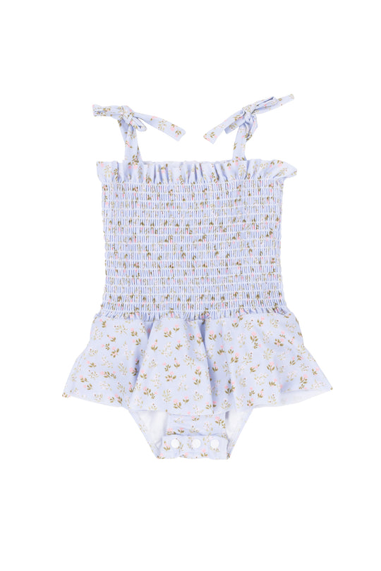 The Ivy City Co Baby Maggie One Piece in Blue Ditsy Floral features shoulder tie straps, a smocked bodice, ruffled peplum skirt, yellow flowers, and crotch snap buttons for easy diaper changes.