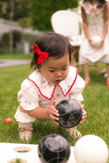 A young child wears the Ivy City Co Baby Maisy Romper in Star Spangled with a red hair bow, crouched on the grass holding a black ball, while another in a similar outfit stands behind, partially visible as they play outside.