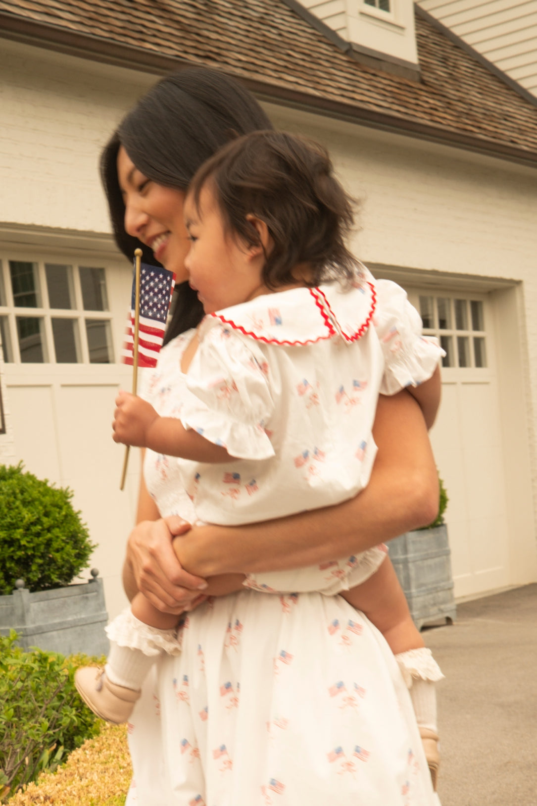 A woman holds a small child outside a white house. Both wear matching white outfits with tiny American flag prints. The child wears the Ivy City Co Baby Maisy Romper in Star Spangled, holding an American flag and leaning toward the woman.