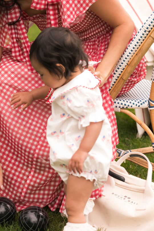 A woman in a red gingham dress helps a toddler wearing the Ivy City Co Baby Maisy Romper in Star Spangled stand on the grass near a cream bag and black bocce balls at a laid-back outdoor gathering.
