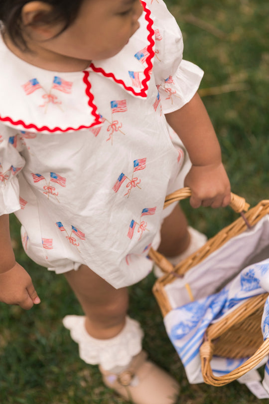 A young child in the Ivy City Co Baby Maisy Romper in Star Spangled, featuring American flag prints and red trim, holds a wicker basket lined with blue and white cloth while standing on grass.