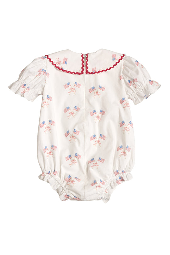 The Ivy City Co Baby Maisy Romper in Star Spangled features puffed short sleeves, red collar trim, tiny American flags and red bows, a scalloped back edge, and elastic leg openings—perfect for your baby girl's Fourth of July outfit.