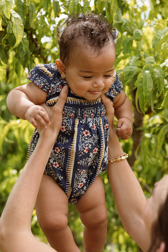 A smiling baby wearing the Ivy City Co Baby Marlow Romper in Navy Fleur is held up in the air by an adult in a garden with green foliage in the background.