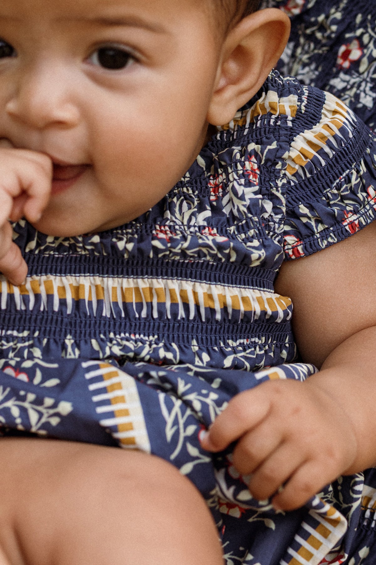 A baby wearing the Ivy City Co Baby Marlow Romper in Navy Fleur—adorned with a charming navy floral design and yellow and white stripes—sits with a finger in their mouth, gazing up and smiling softly.