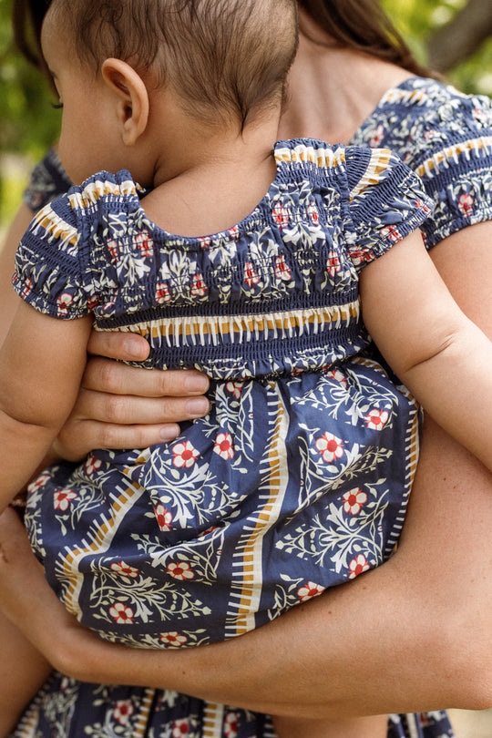 An adult holds a baby dressed in the Ivy City Co Baby Marlow Romper in Navy Fleur, both partially visible outdoors with blurred greenery in the background.