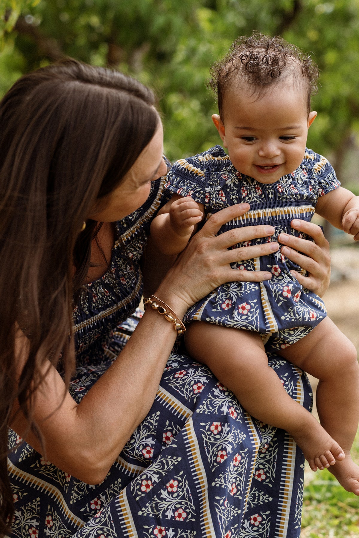 A woman holds a smiling baby outdoors, both dressed in matching blue floral outfits. The little one wears the charming Baby Marlow Romper in Navy Fleur by Ivy City Co. Lush greenery blurs softly in the background, suggesting a natural setting.