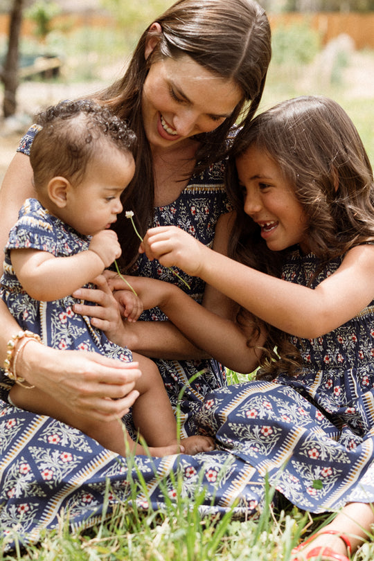 A woman and two young children sit on the grass, all dressed in matching blue patterned outfits from Ivy City Co. The baby looks adorable in the Baby Marlow Romper in Navy Fleur, as the older child playfully holds a small flower nearby and they all smile together.