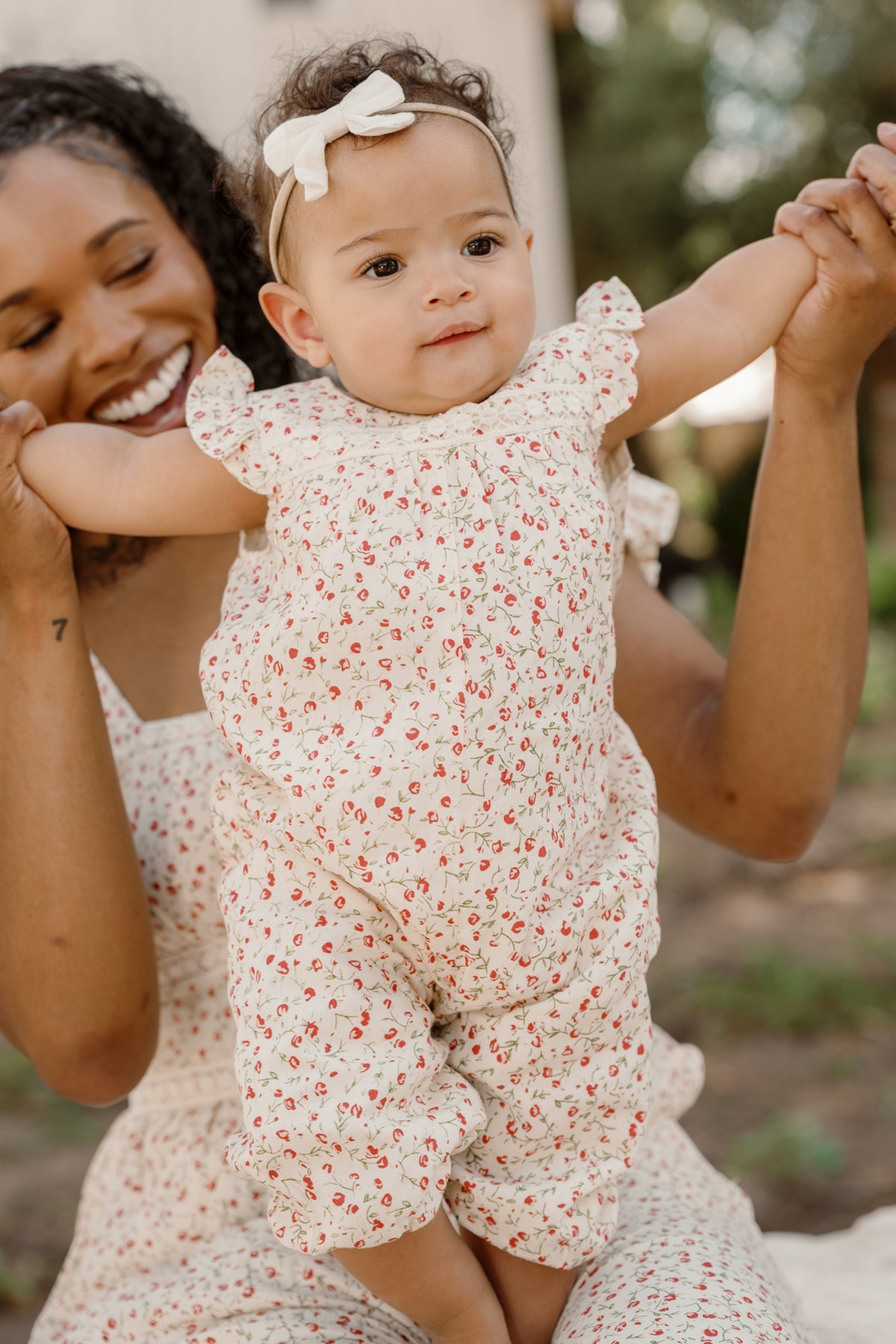 A smiling woman holds up a baby dressed in the Ivy City Co Baby Mirabelle Romper in Cream Ditsy Floral, featuring delicate lace trim and a matching headband. Both are outside, and the baby has arms raised, looking curious and happy.