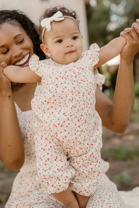 A smiling woman holds up a baby dressed in the Ivy City Co Baby Mirabelle Romper in Cream Ditsy Floral, featuring delicate lace trim and a matching headband. Both are outside, and the baby has arms raised, looking curious and happy.