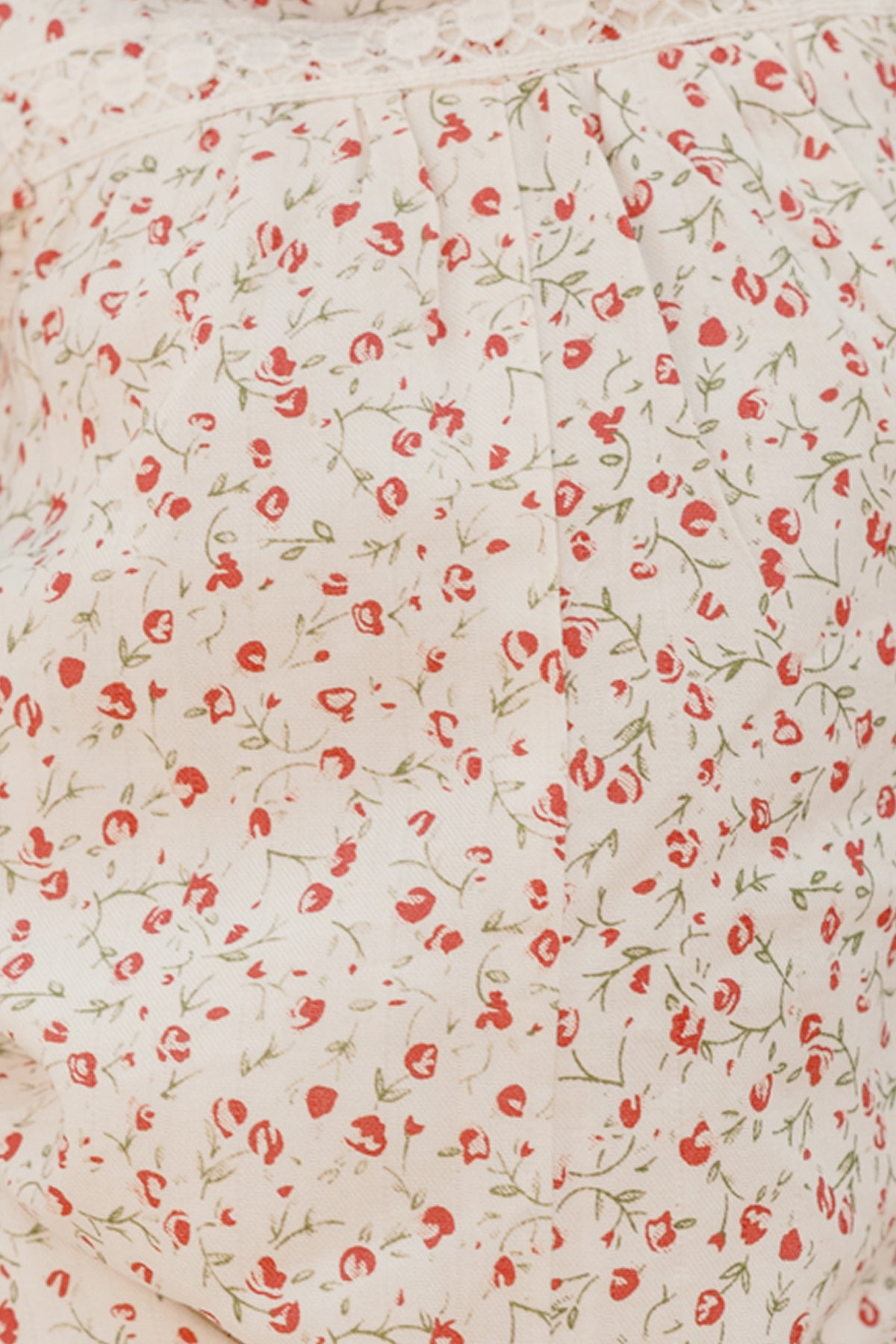 Close-up of the Ivy City Co Baby Mirabelle Romper in Cream Ditsy Floral, featuring light fabric with a delicate red and green floral pattern and lace trim at the top—perfect for crafting a charming baby look. The background color is off-white or cream.