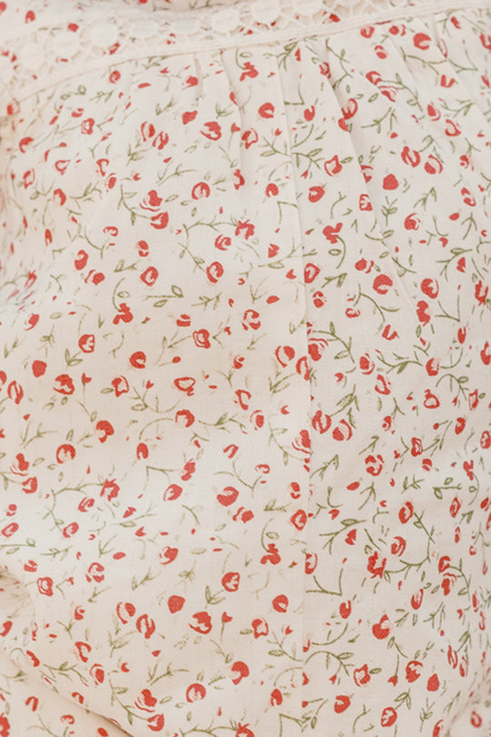Close-up of the Ivy City Co Baby Mirabelle Romper in Cream Ditsy Floral, featuring light fabric with a delicate red and green floral pattern and lace trim at the top—perfect for crafting a charming baby look. The background color is off-white or cream.