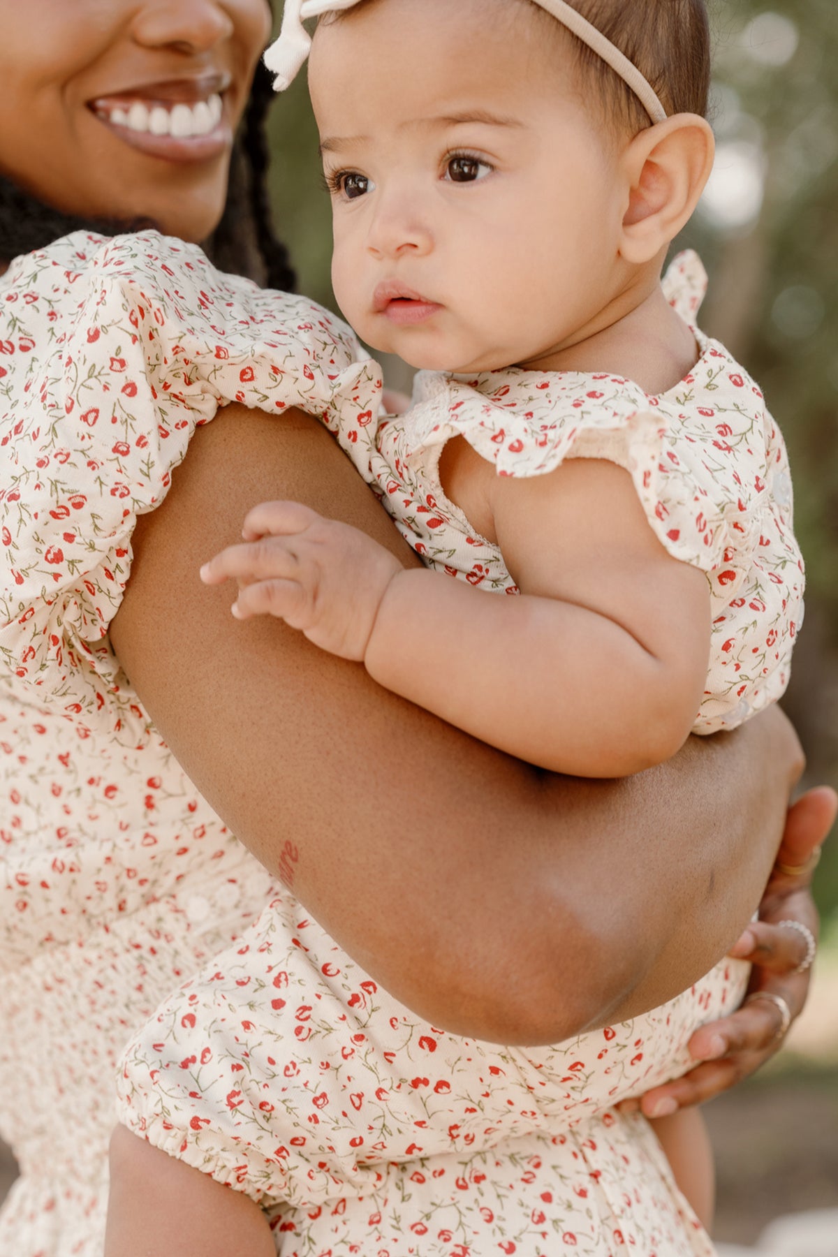 A woman smiles as she holds a baby close. Both are dressed in matching cream outfits adorned with small red floral prints and delicate lace trim. The baby wears the Ivy City Co Baby Mirabelle Romper in Cream Ditsy Floral, looking away with a calm expression.