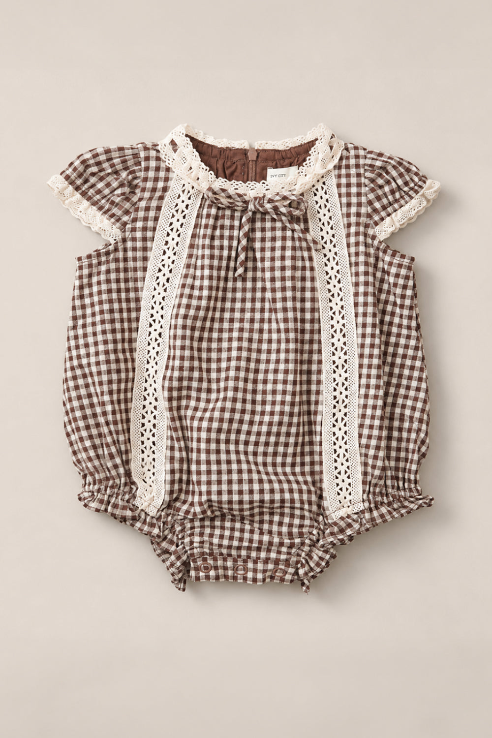 The Ivy City Co Baby Palermo Romper in Hickory Gingham, featuring white lace trim, short puff sleeves, and ruffled leg openings, is laid flat on a neutral background. This charming cotton-blend romper is perfect for any occasion.