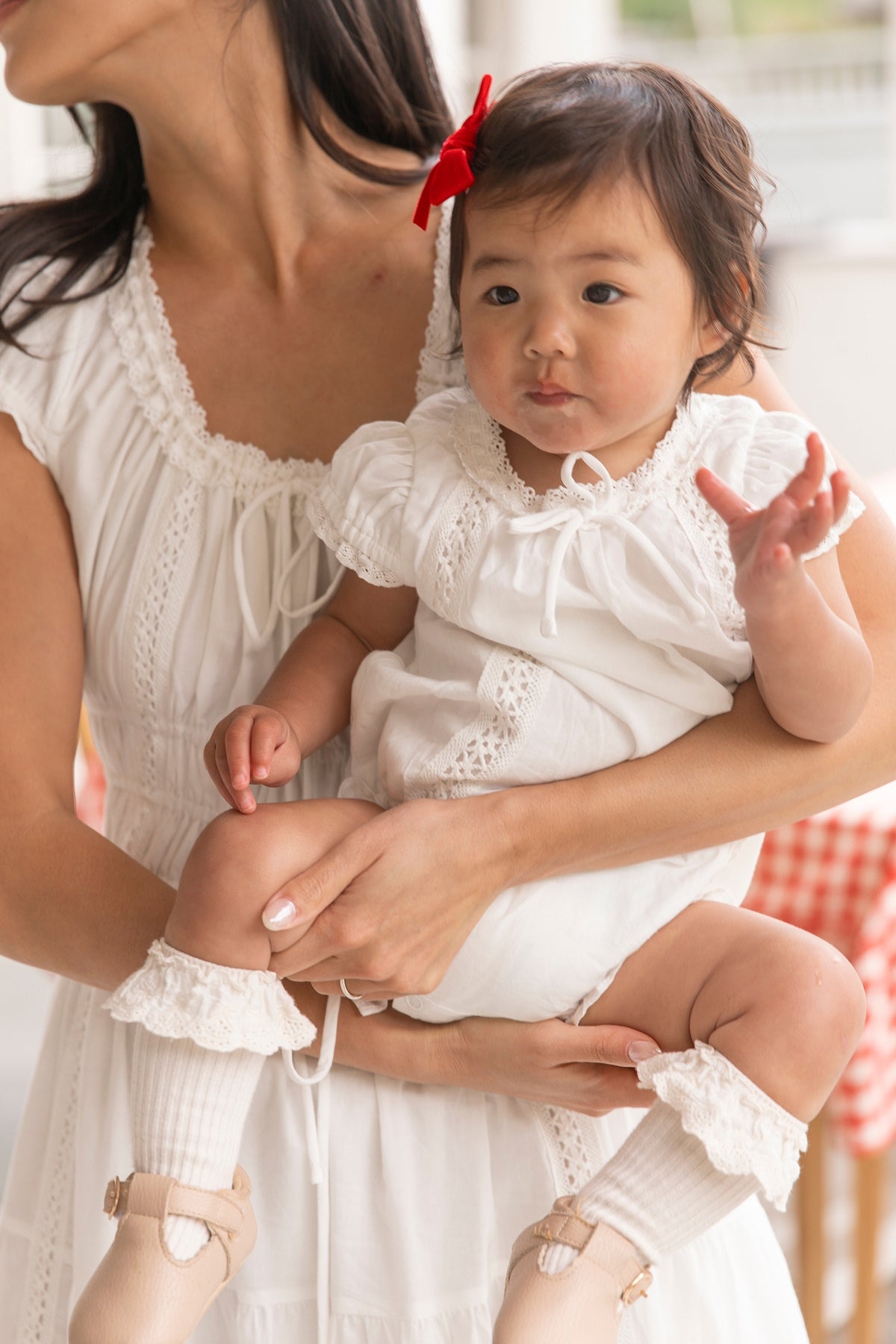 A woman in white holds her baby girl dressed in the Ivy City Co Baby Palermo Romper in White, accessorized with frilly socks, beige shoes, and a red bow—the perfect matching moment for mom and daughter.