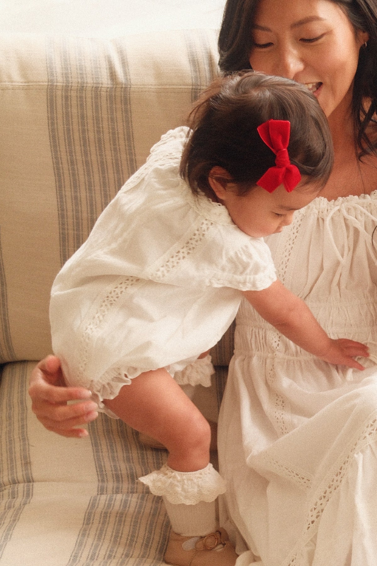 A woman in a white dress smiles at a toddler wearing the Ivy City Co Baby Palermo Romper in White with a red bow, as the child climbs onto her lap for a sweet Mom and Me matching moment.