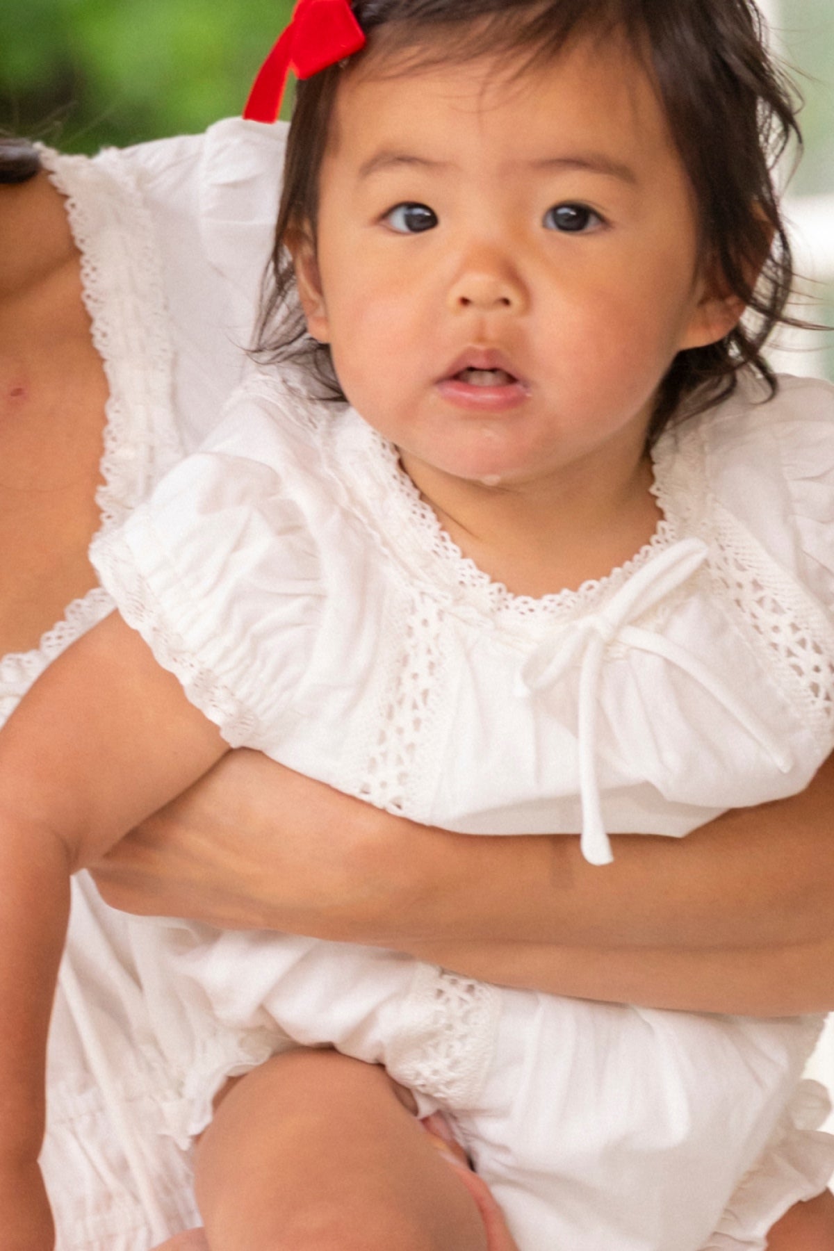A young child with a red bow and short dark hair is held by an adult, wearing the Ivy City Co Baby Palermo Romper in White with ruffled sleeves and lace accents, perfect for Mom and Me matching, while looking slightly off camera.