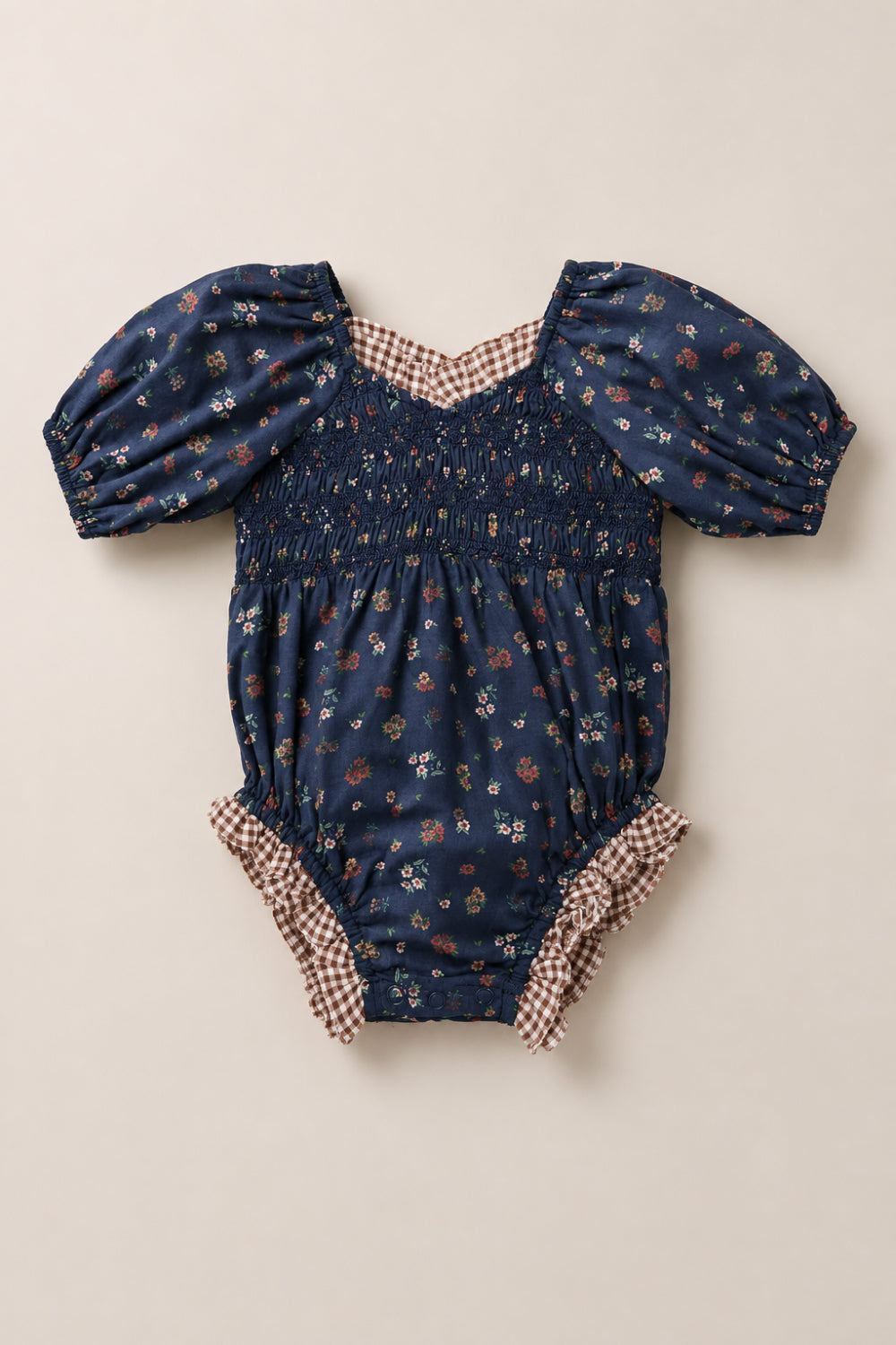 The Baby Roselyn Romper in Midnight Lovelorn Ditsy by Ivy City Co features a navy blue base adorned with a delicate small floral print, short puff sleeves, a smocked bodice, and brown gingham trim along the neckline and leg openings. Laid flat on a light background, this charming cotton romper combines comfort and style for your little one.
