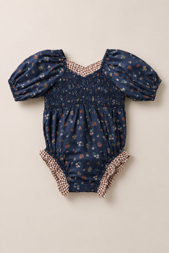The Baby Roselyn Romper in Midnight Lovelorn Ditsy by Ivy City Co features a navy blue base adorned with a delicate small floral print, short puff sleeves, a smocked bodice, and brown gingham trim along the neckline and leg openings. Laid flat on a light background, this charming cotton romper combines comfort and style for your little one.