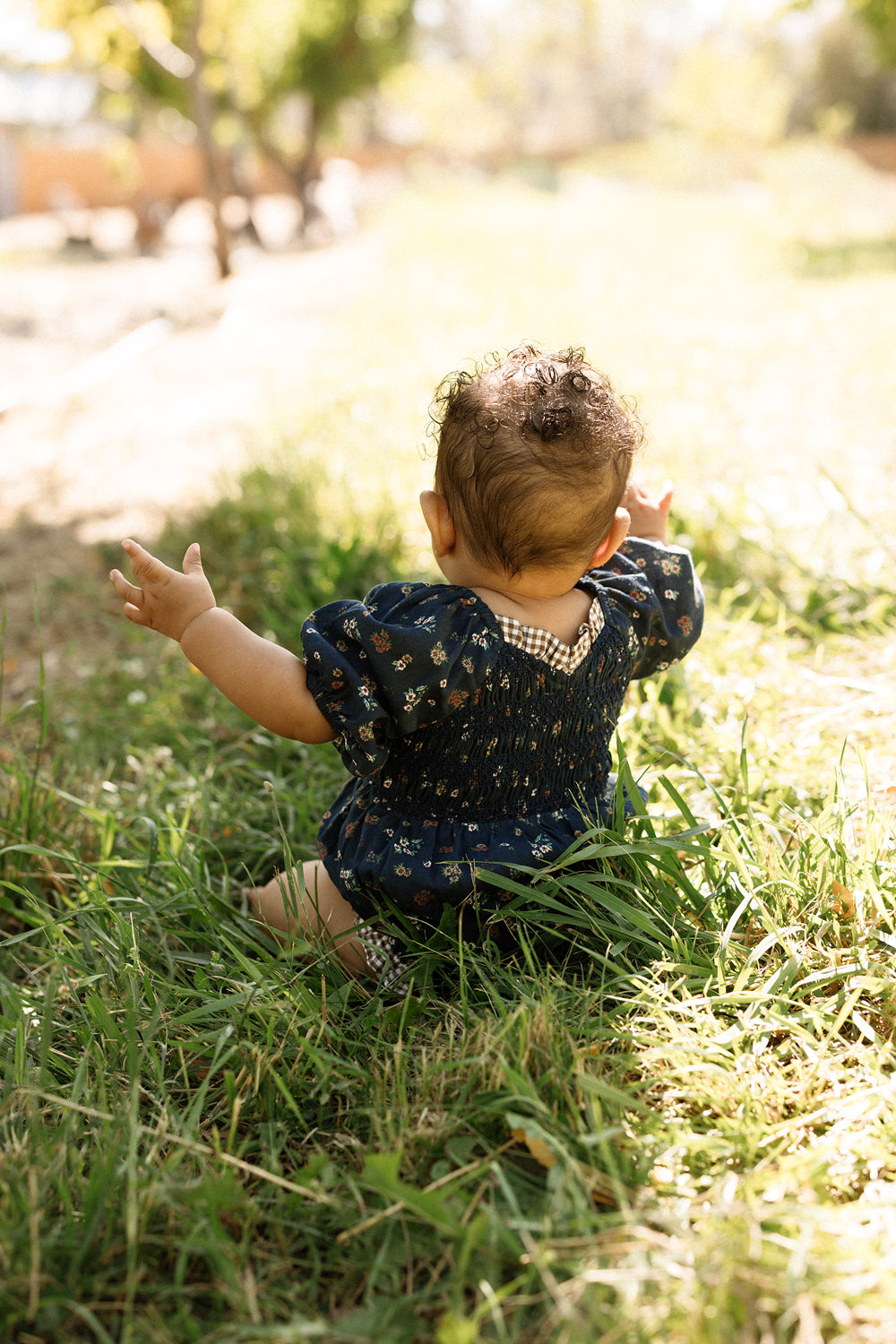 A baby with curly hair sits in sunlit grass, seen from behind, wearing the Ivy City Co Baby Roselyn Romper in Midnight Lovelorn Ditsy, its smocked bodice visible as their arms reach toward a sun-dappled park.