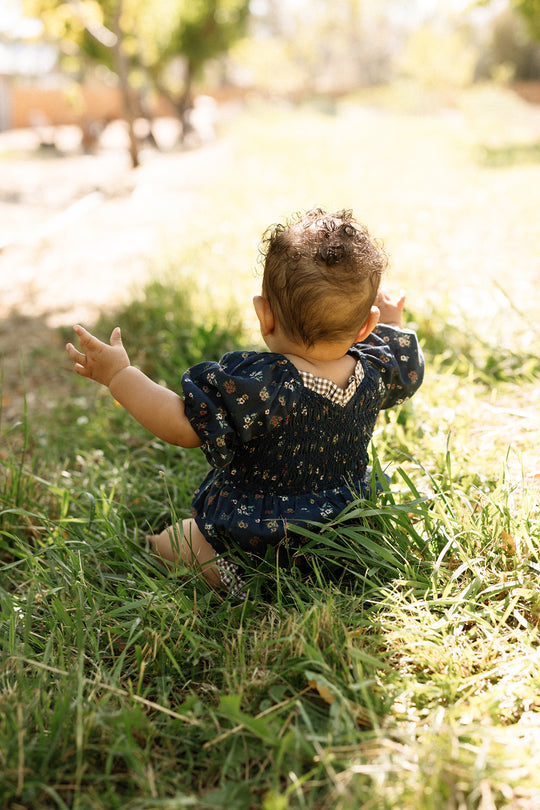A baby with curly hair sits in sunlit grass, seen from behind, wearing the Ivy City Co Baby Roselyn Romper in Midnight Lovelorn Ditsy, its smocked bodice visible as their arms reach toward a sun-dappled park.