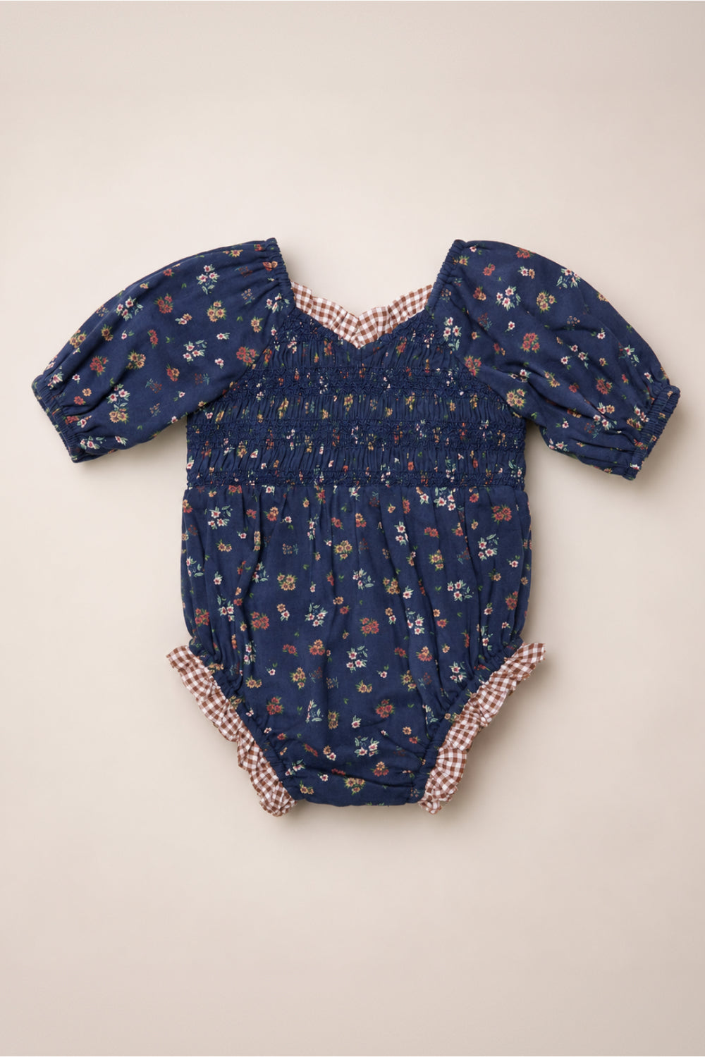 The Baby Roselyn Romper in Midnight Lovelorn Ditsy by Ivy City Co features a navy blue floral pattern, puffed short sleeves, a smocked bodice, and red gingham trim at the collar and leg openings. Crafted from soft cotton, it is shown laid flat on a light background.