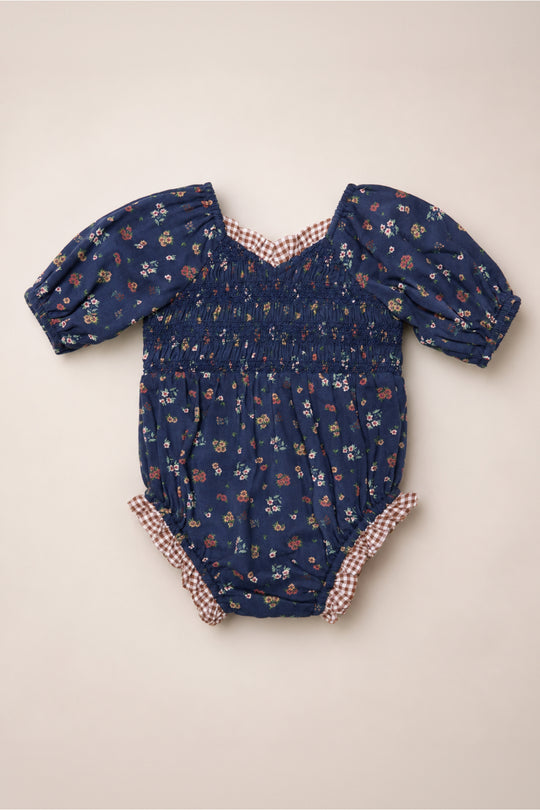 The Baby Roselyn Romper in Midnight Lovelorn Ditsy by Ivy City Co features a navy blue floral pattern, puffed short sleeves, a smocked bodice, and red gingham trim at the collar and leg openings. Crafted from soft cotton, it is shown laid flat on a light background.