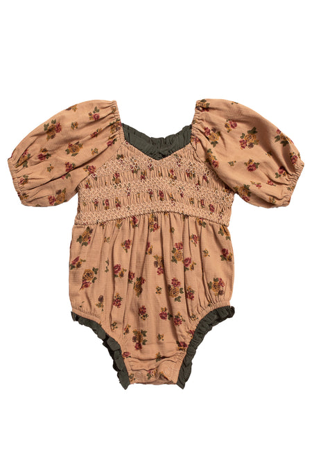 The Ivy City Co Baby Roselyn Romper in Desert Blossom features puffed short sleeves, a smocked bodice, pinkish-tan cotton with a small floral print, and green ruffled trim at the neckline and leg openings.