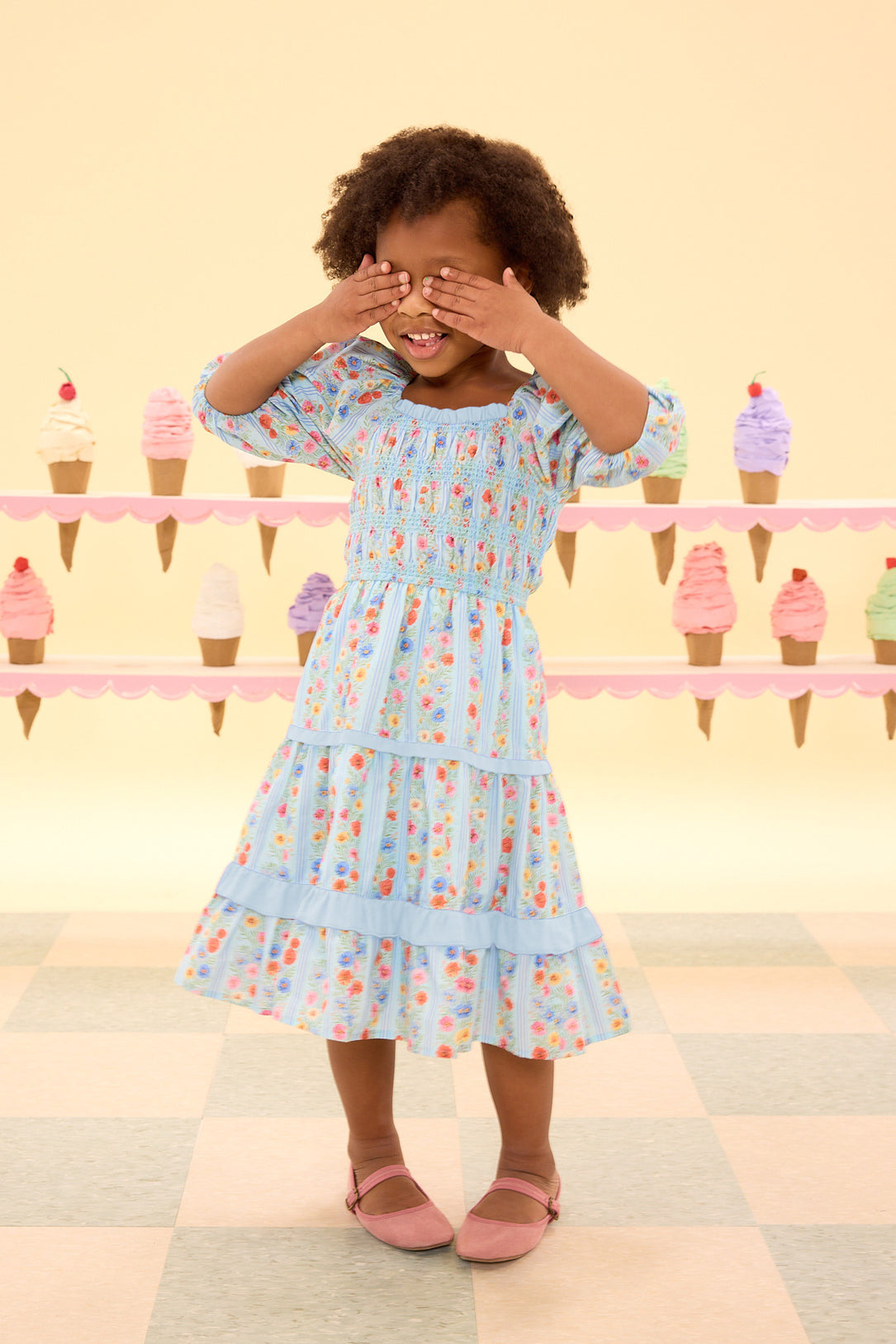 Young girl in a light blue dress with ice cream pattern covering her eyes against a pastel background with ice cream cones.