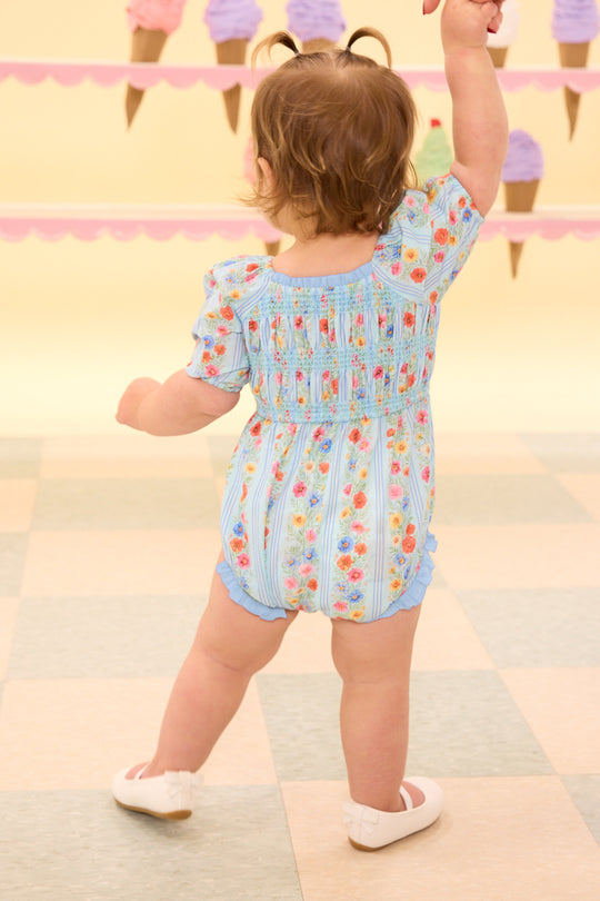 the back of a baby wearing a blue floral baby romper standing on the a tiled floor in front of an ice cream wall