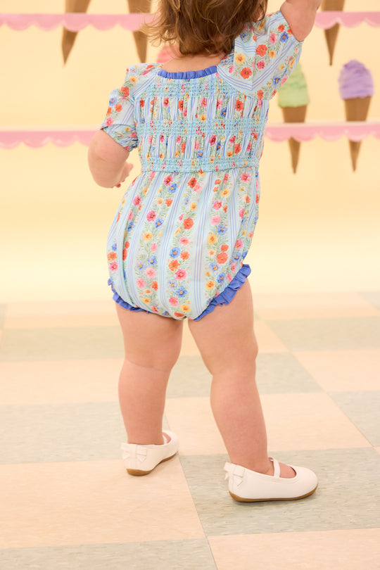 Child wearing a colorful floral romper on a patterned floor.