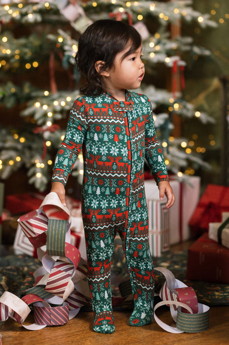 A young child wearing the Ivy City Co Baby So Soft Onesie in Alpine Noel holds a paper garland, standing before a decorated Christmas tree with gifts underneath.