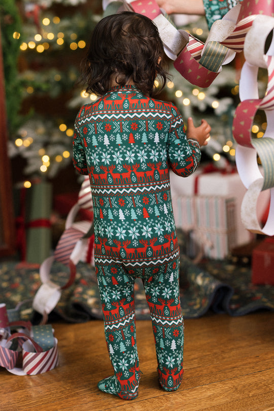 A child in the Ivy City Co Baby So Soft Onesie in Alpine Noel stands on a wooden floor, surrounded by paper chains and holiday gifts near a glowing Christmas tree.