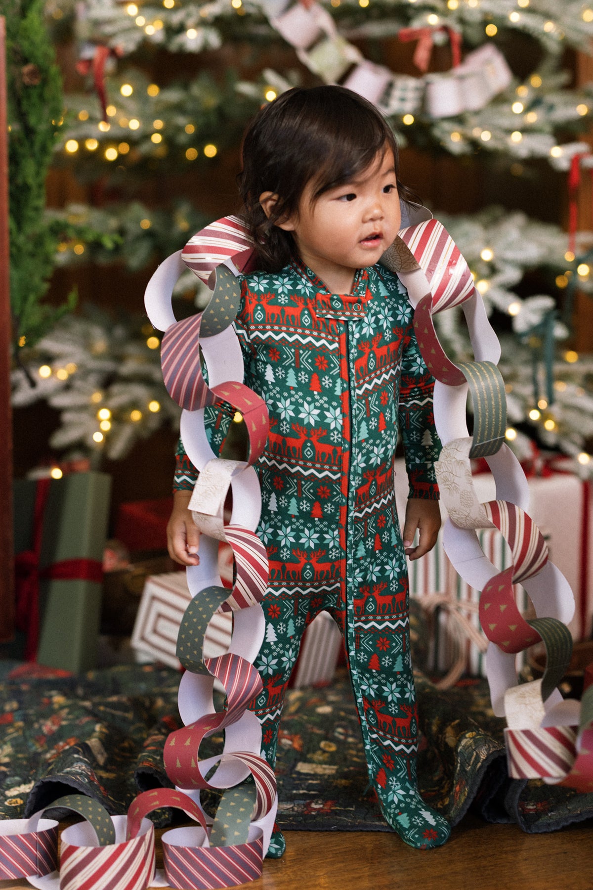 A toddler in the Ivy City Co Baby So Soft Onesie in Alpine Noel stands by a Christmas tree, draped with a festive paper chain garland. Wrapped gifts and twinkling lights glow in the background.