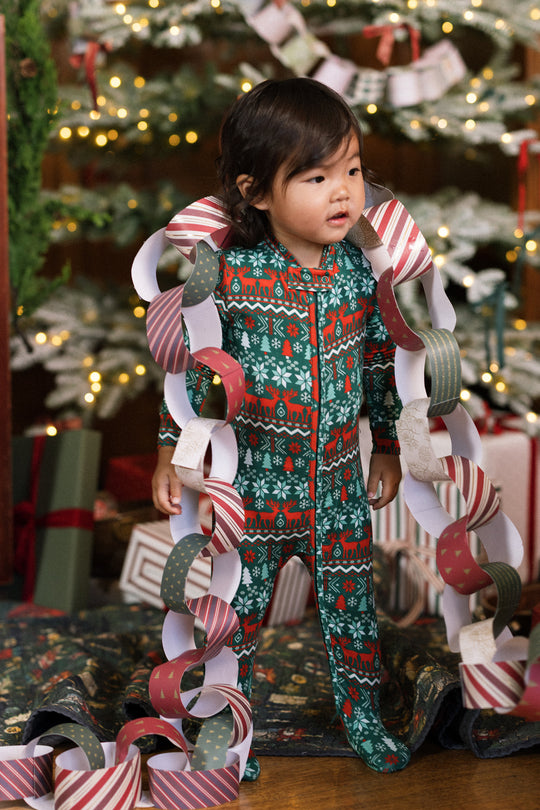 A toddler in the Ivy City Co Baby So Soft Onesie in Alpine Noel stands by a Christmas tree, draped with a festive paper chain garland. Wrapped gifts and twinkling lights glow in the background.
