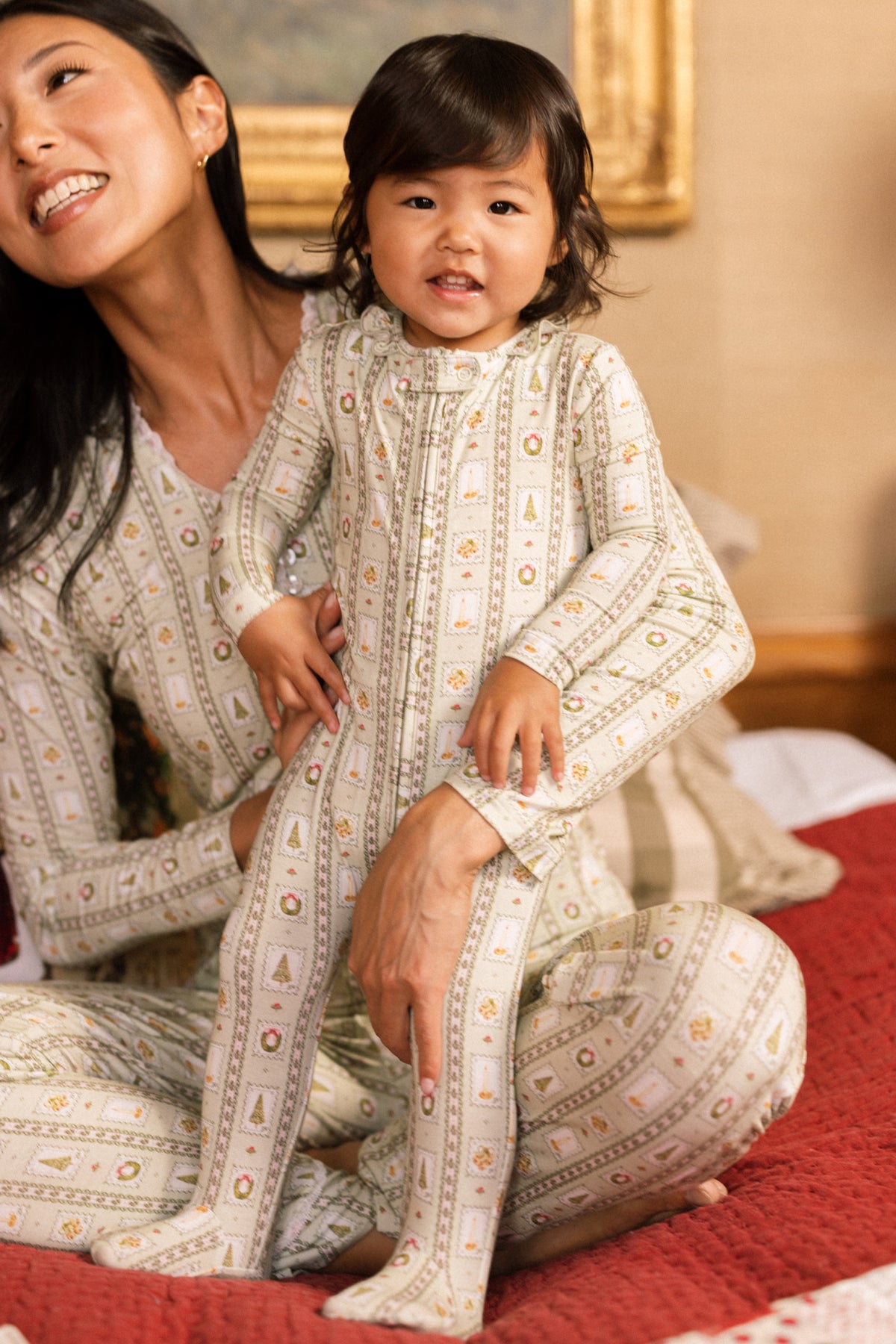 A smiling toddler in the Ivy City Co Baby So Soft Onesie in Merry Mail Linear sits on a bed, held by a woman in matching family pajamas, with a picture frame hanging on the wall behind them.