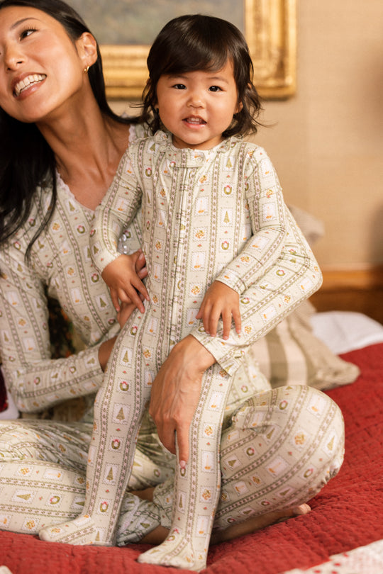 A smiling toddler in the Ivy City Co Baby So Soft Onesie in Merry Mail Linear sits on a bed, held by a woman in matching family pajamas, with a picture frame hanging on the wall behind them.