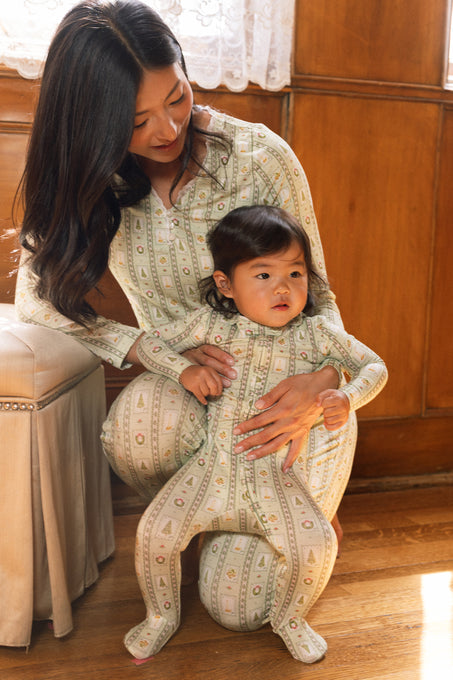 A woman kneels on the floor, smiling at a child in her lap, both cozy in Ivy City Co’s Baby So Soft Onesie in Merry Mail Linear. Sunlight pours in, highlighting their warmth and joy.