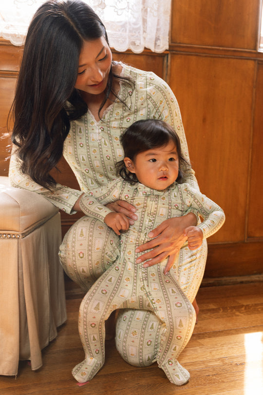 A woman kneels on the floor, smiling at a child in her lap, both cozy in Ivy City Co’s Baby So Soft Onesie in Merry Mail Linear. Sunlight pours in, highlighting their warmth and joy.