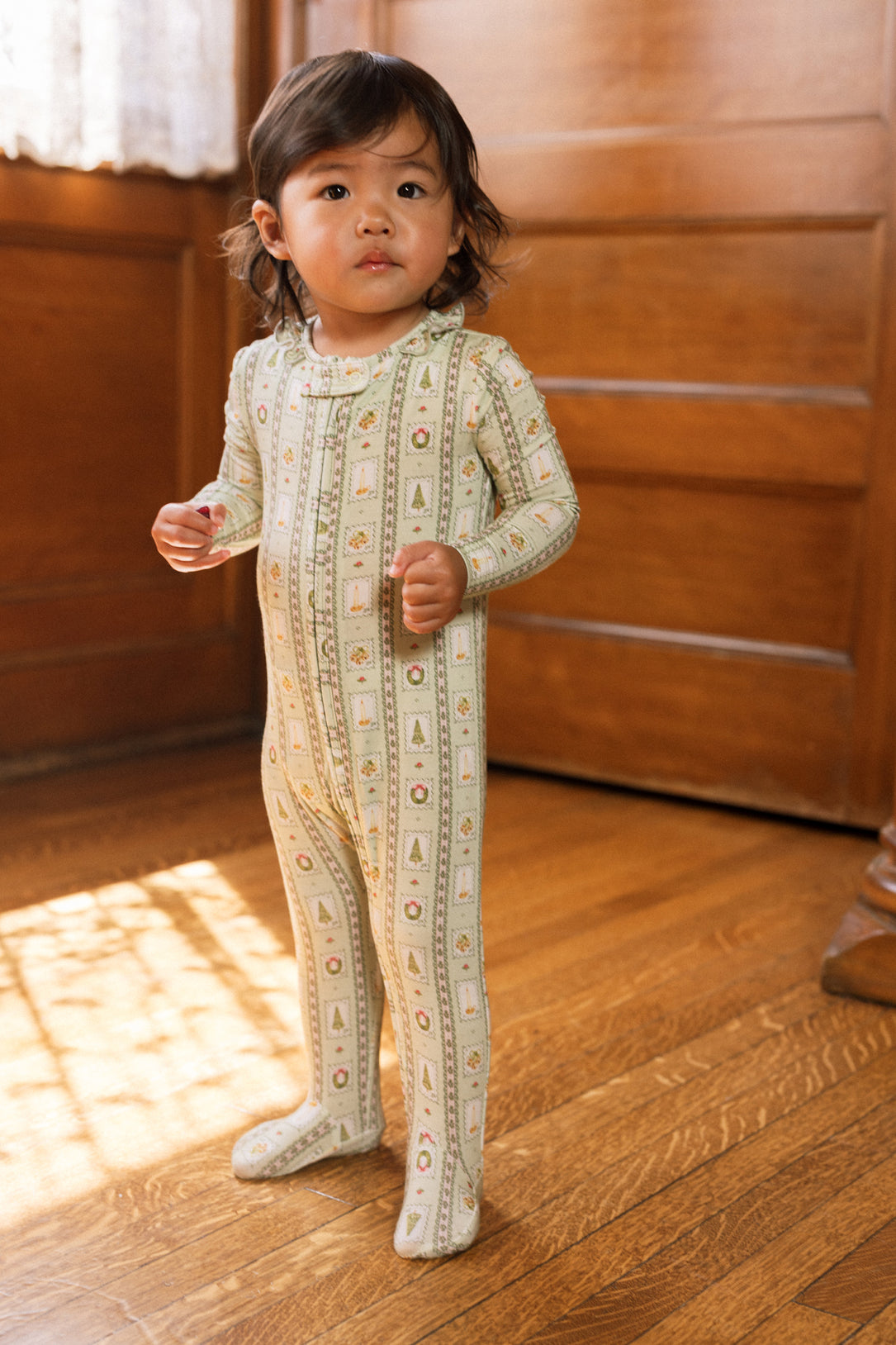 A young child with medium-length dark hair stands on a wooden floor, wearing the Ivy City Co Baby So Soft Onesie in Merry Mail Linear. Warm light streams in from the left.