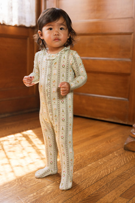 A young child with medium-length dark hair stands on a wooden floor, wearing the Ivy City Co Baby So Soft Onesie in Merry Mail Linear. Warm light streams in from the left.