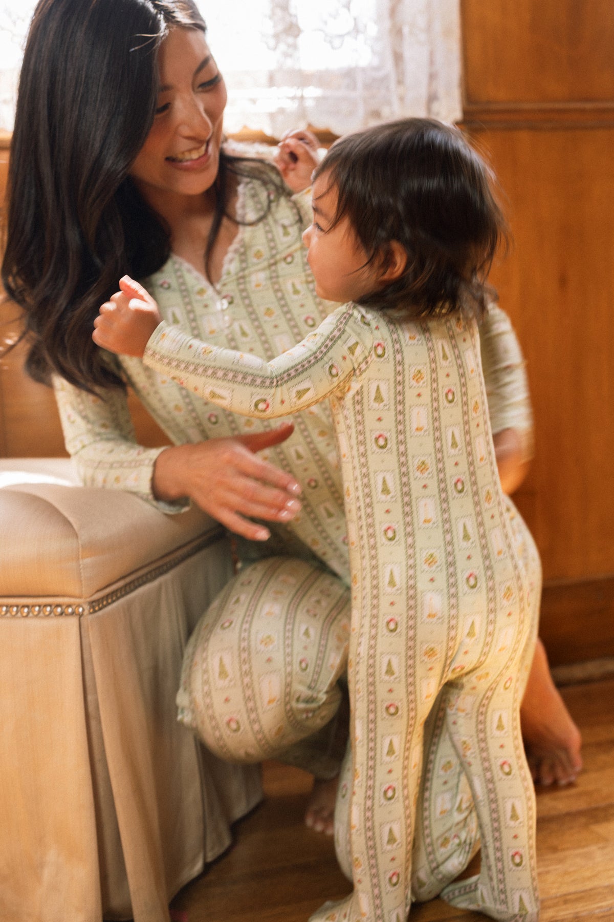 A woman kneels, smiling at a young child wearing the Ivy City Co Baby So Soft Onesie in Merry Mail Linear, both basking in warm light by a lace-curtained window.