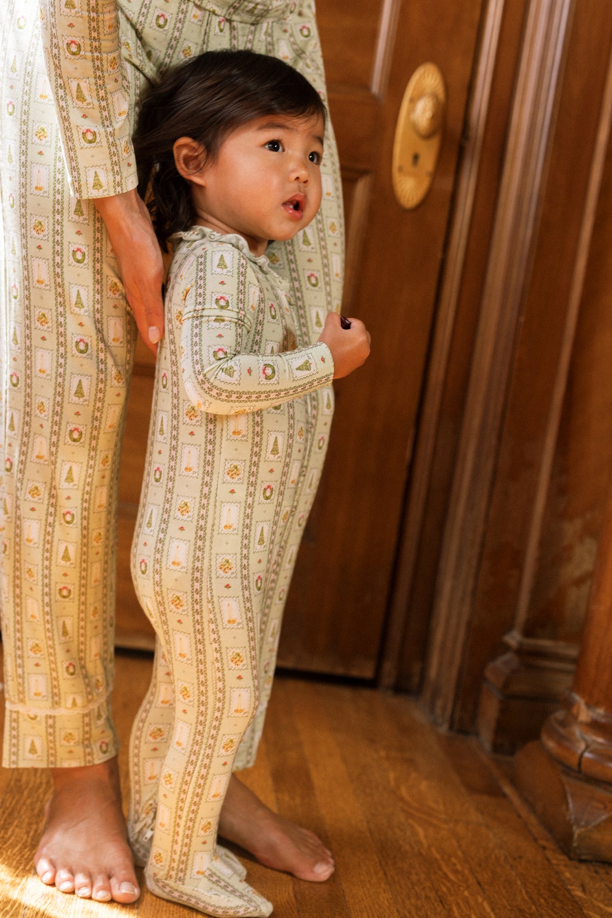 A toddler in patterned pajamas stands on a wooden floor, holding an adult’s hand. Both wear matching Ivy City Co pj’s; the Baby So Soft Onesie in Merry Mail Linear stands out by the wooden door with a brass handle.