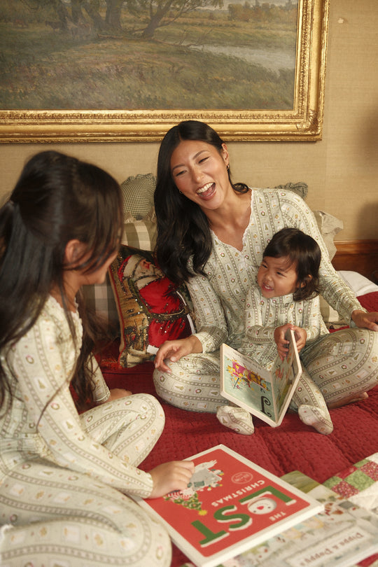 A smiling woman and two children in Baby So Soft Onesie in Merry Mail Linear by Ivy City Co sit on a bed reading Christmas books, surrounded by festive decor.