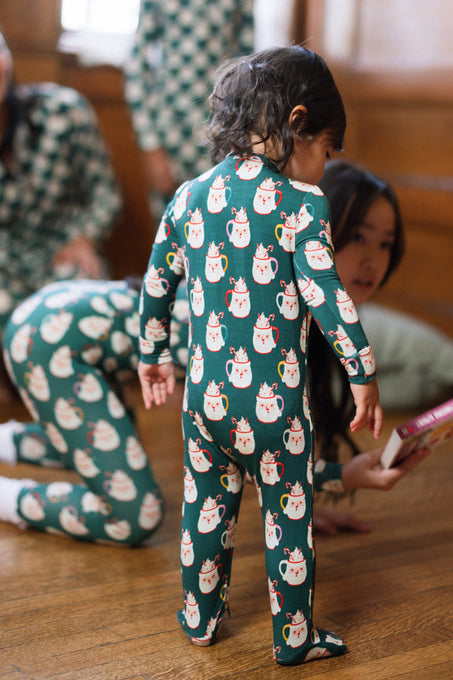 A toddler wearing the Baby So Soft Onesie in Very Merry Santa by Ivy City Co stands on a wooden floor, while adults in matching cotton-modal knit pajamas sit and interact in the background.